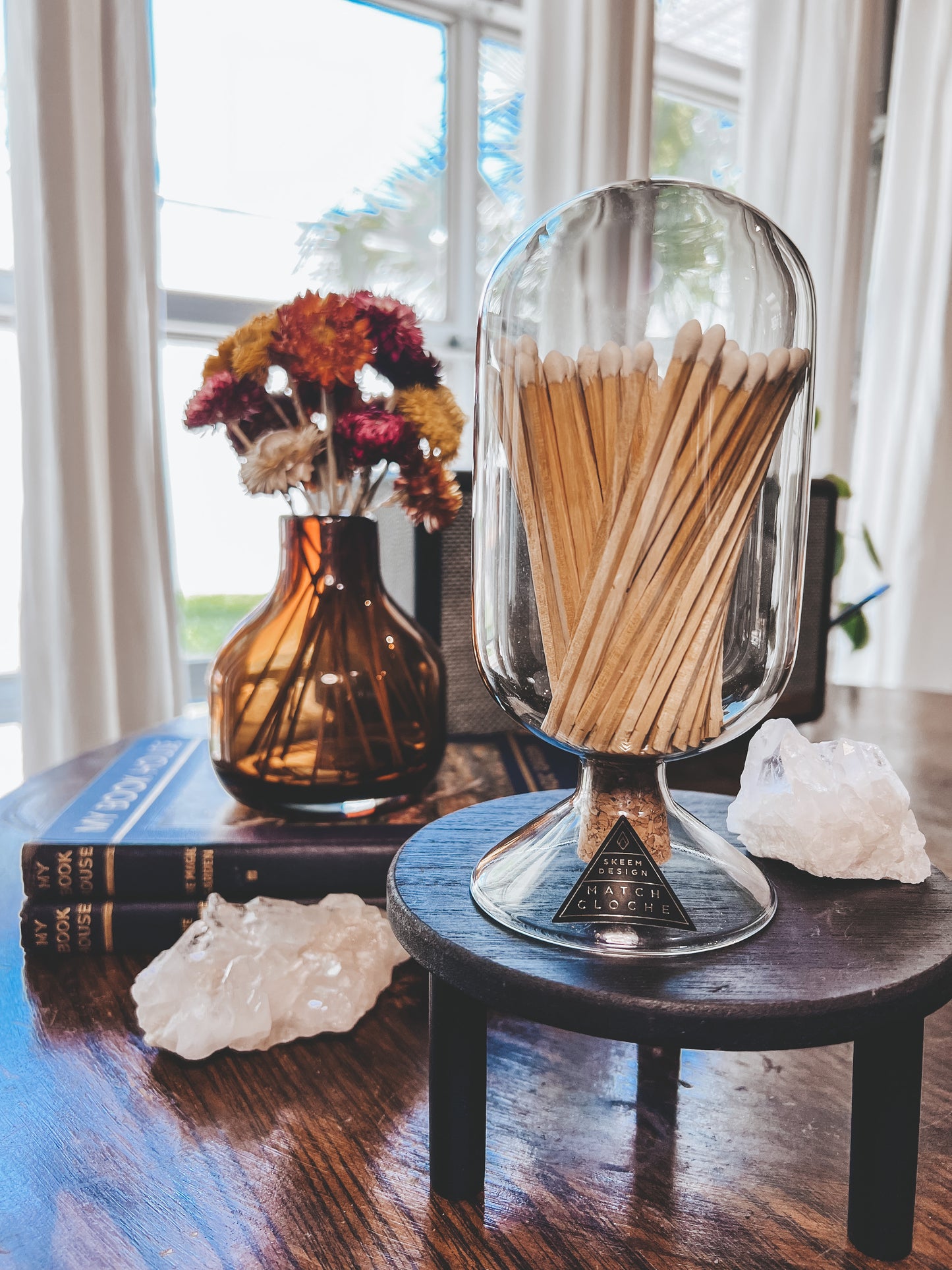 Skeem Designs match cloche in Smoke color sitting upon a dark wooden pedestal with a palm sized quartz stone beside it. Behind the cloche is a small amber colored vase filled with spring colored flowers. Vase is sitting on top of two brown books. Scene is displayed on wooded table.