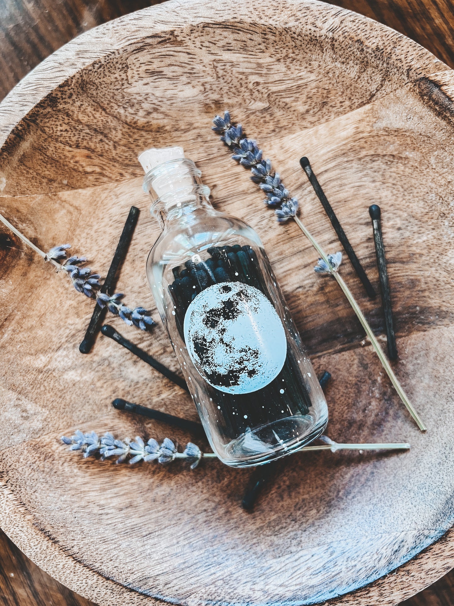 Miniture match bottle that is made of clear glass with a cork lid. Bottle has a white silk screened image of a full moon and all its craters printed on it. The match bottle is resting on a light colored wooded bowl with sprigs of dried lavender spread around it. The match sticks are black wood with black tips.