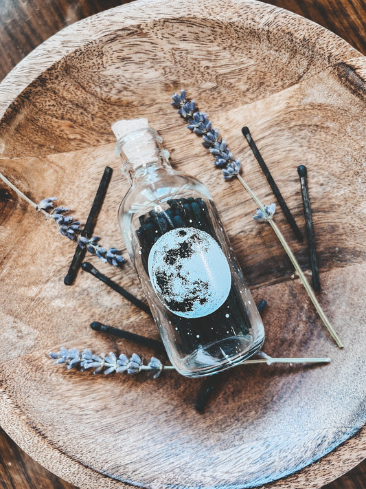 Miniture match bottle that is made of clear glass with a cork lid. Bottle has a white silk screened image of a full moon and all its craters printed on it. The match bottle is resting on a light colored wooded bowl with sprigs of dried lavender spread around it. The match sticks are black wood with black tips.