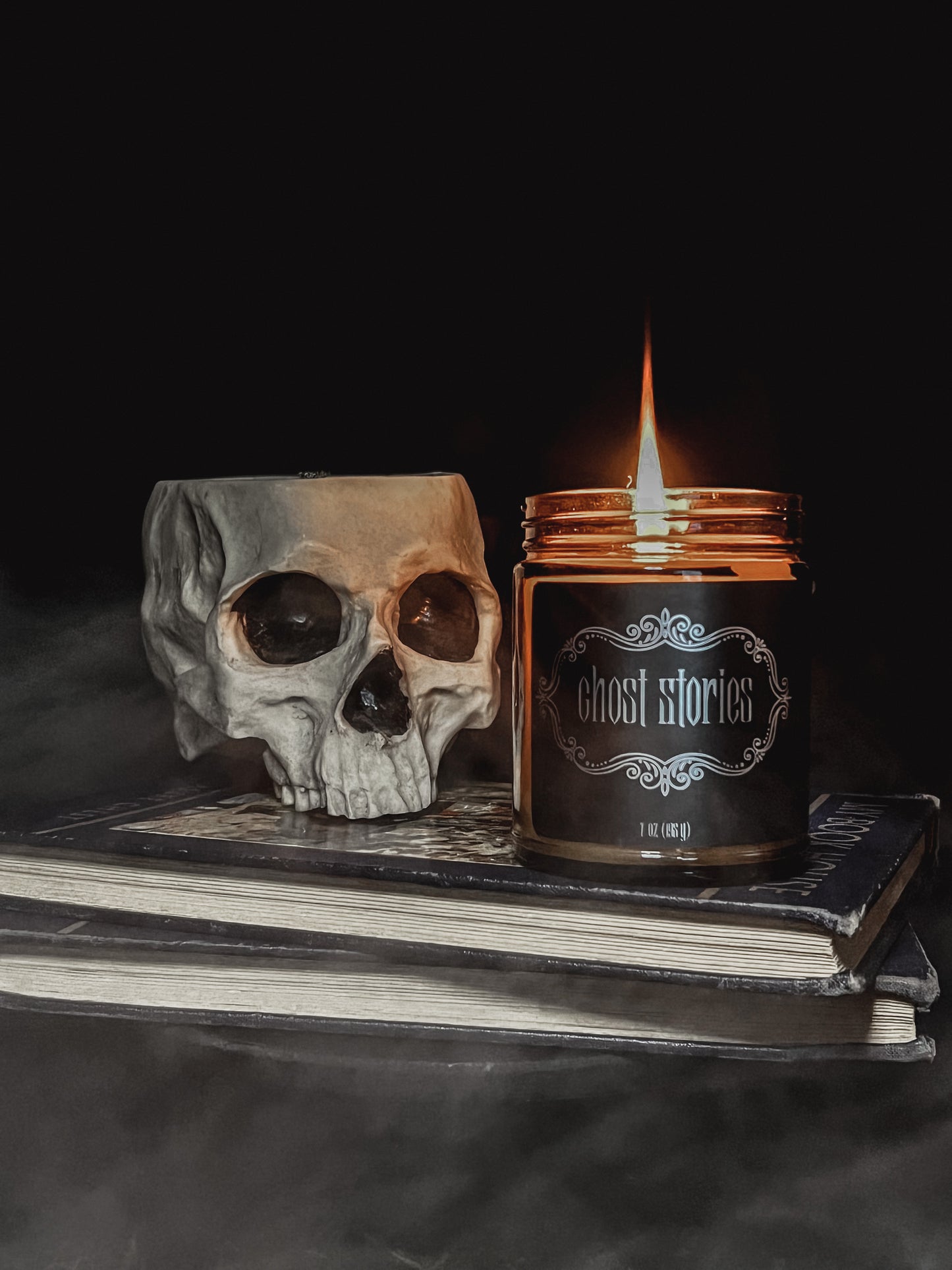 Halloween amber glass jar candle with black label embellished with metallic filigree and the text "Ghost Stories". Candle is lit and sitting on top of two hardback books with black covers. Beside the candle is a creepy grey colored skull with black eye sockets. Top of the skull is open.