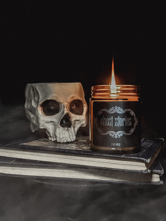 Halloween amber glass jar candle with black label embellished with metallic filigree and the text "Ghost Stories". Candle is lit and sitting on top of two hardback books with black covers. Beside the candle is a creepy grey colored skull with black eye sockets. Top of the skull is open.