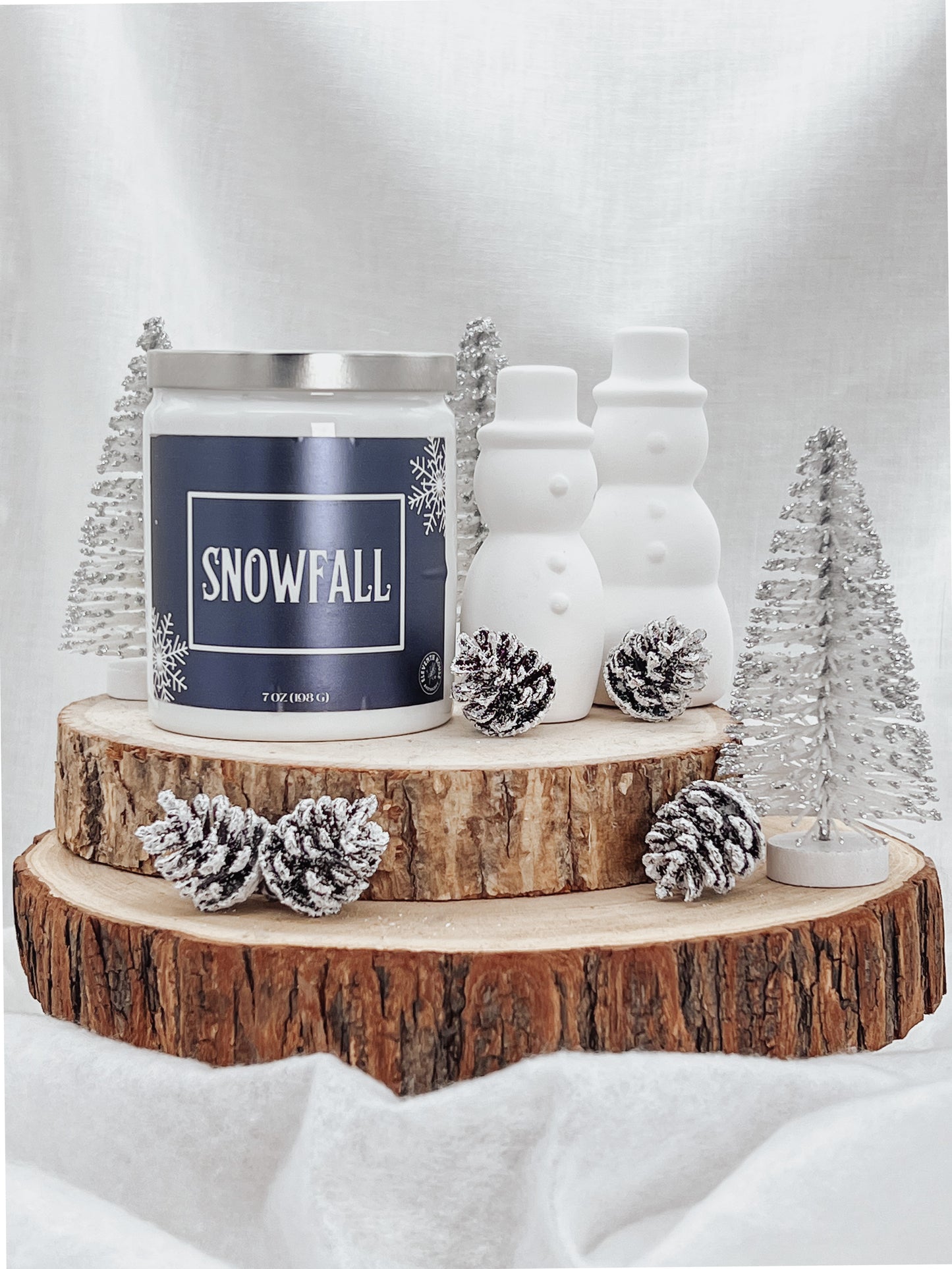 A white jar candle with a silver metal lid and a blue metallic label with images of white snow flakes with the text "Snowfall" printed on it. It sits on top of two medium sized tree stump discs. Placed along side it are two white snowmen and a few snow covered pine cones. Place with the pine cones are mini snow covered trees.