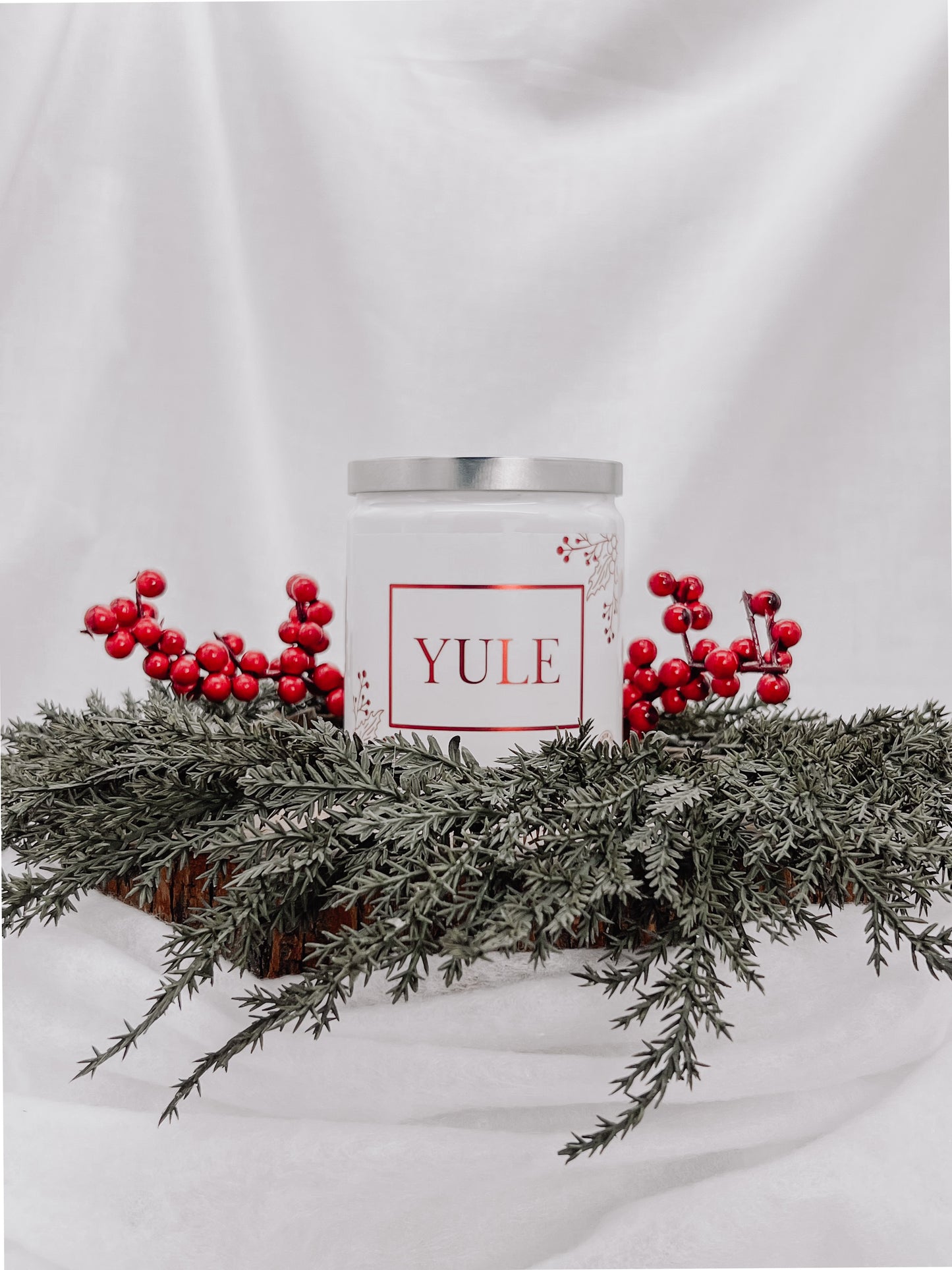 A white jar candle with a silver metal lid and a white metallic label with images of red berries attached to a branch with the text "Yule" printed in red. Candle sits on top of one medium sized tree stump disc surrounded by evergreen branches and red berries. Background is a white back drop.