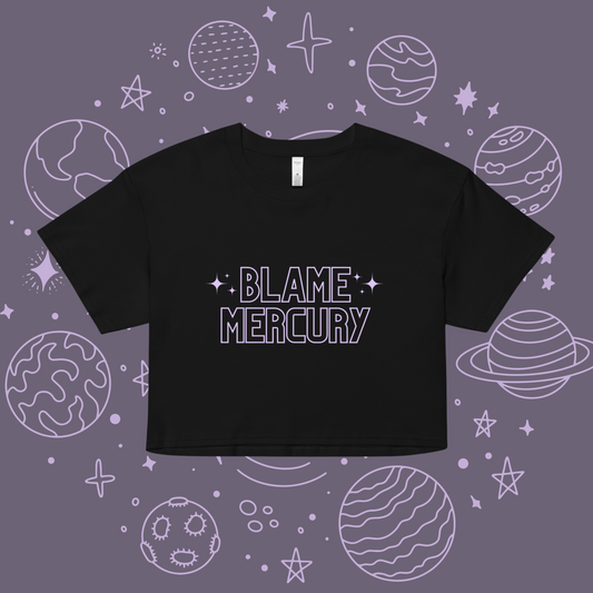 This is a typography style crop top with the caption "Blame Mercury" in a light purplish gray color font. Each letter of the caption is outlined and not filled with this solid color. The word "Blame" is stacked on top of the word "Mercury". On each side of the word "Blame" there are three stars that are clustered together. This design is printed on a black colored top or shirt. .