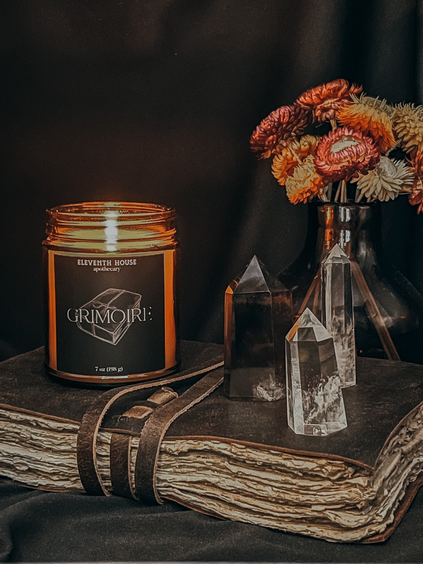 Amber jar candle with a black matte style label including the text "Grimoire" and an image of old world books printed in white. Candle is lit and sitting on leather bound hand made style book. Sitting to the left of the candle on top of the book are three small crystal towers. Behind the towers is a small amber colored vase filled with spring colored flowers. Vase is sitting on top of the book.