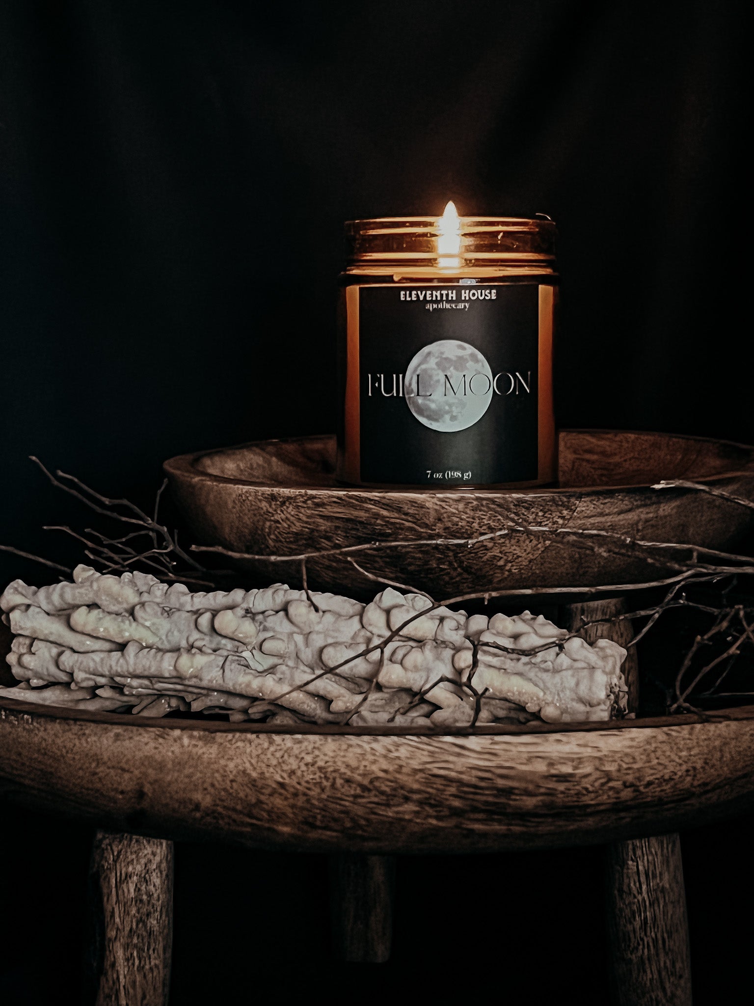 Amber jar candle with a black matte style label including the text "FULL MOON" and an image of a full moon printed in white. Candle is lit sitting on top a small wooden pedestal. Accompanied by a crystalized piece of wood that is stacked on top a larger wooden pedestal.