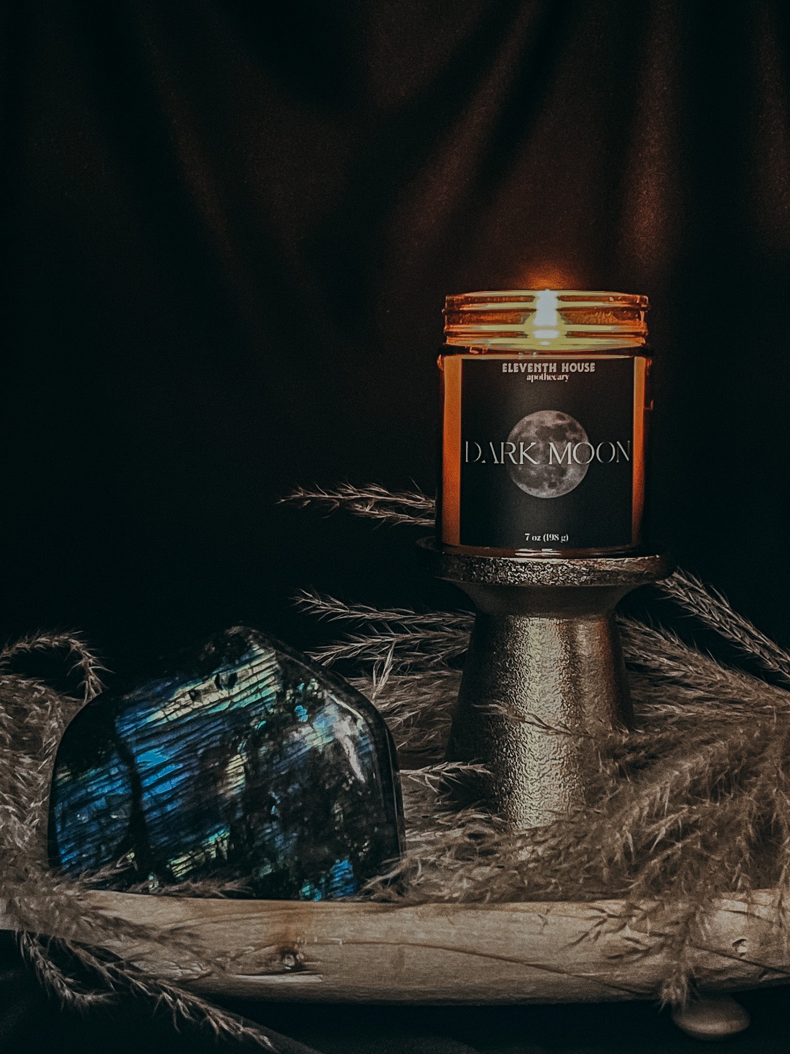 Amber jar candle with a black matte style label including the text "DARK MOON" and an image of a dark moon printed in white lit on a golden pedistal. Surrounded by wild hand picked greenery accompanied by a large glistening labradorite crystal place on wooden plate.