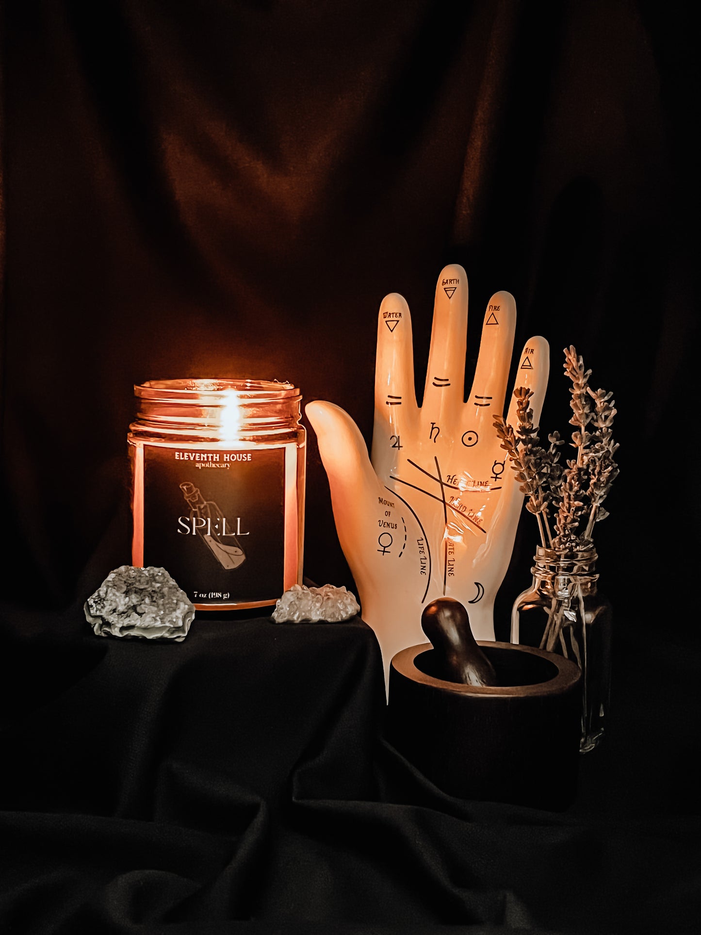 Amber jar candle with a black matte style label including the text "Spell" with an image of potion bottle printed in white. Candle is lit and sitting on black riser beside a ceramic palmistry hand, mortar and pestle, and sprigs of dried lavender. Two quartz crystals accompany the candle.