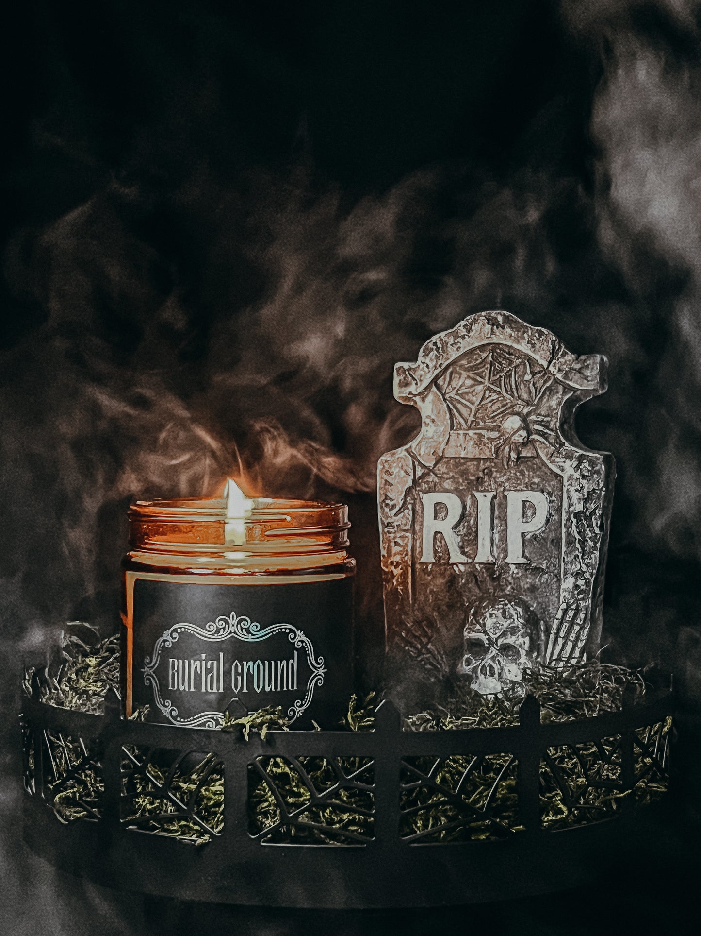 Halloween amber glass jar candle with black label embellished with metallic filigree and the text "Burial Ground". Surrounded with graveyard scene and headstone with RIP and skull printed on it. Scene is contained within a circular metal fence made of spider webs.