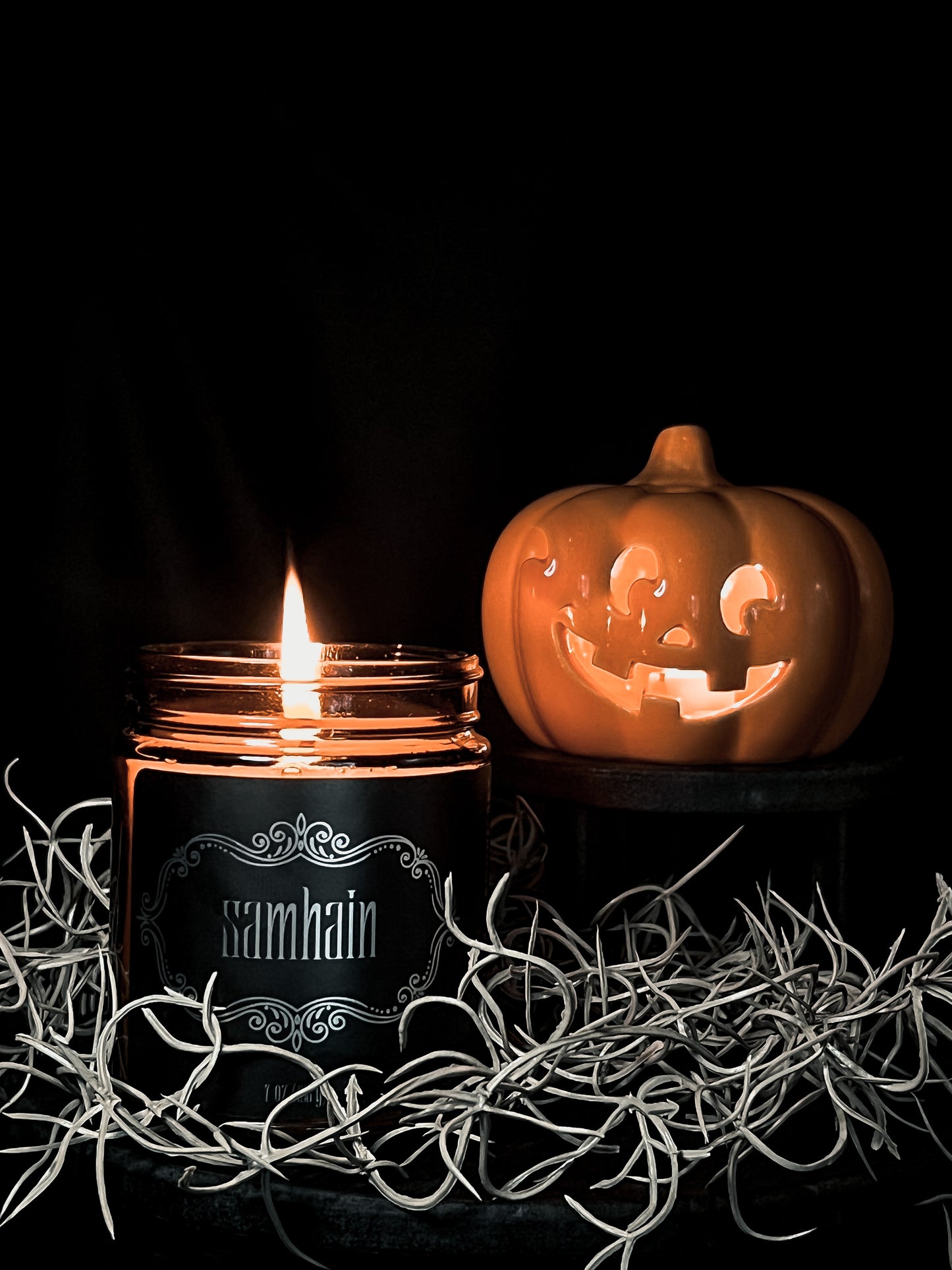 Halloween amber glass jar candle with black label embellished with metallic filigree and the text "Samhain". Wiry sticks surround the scene with a small jack-o-lantern displayed beside the candle. Background is solid black.