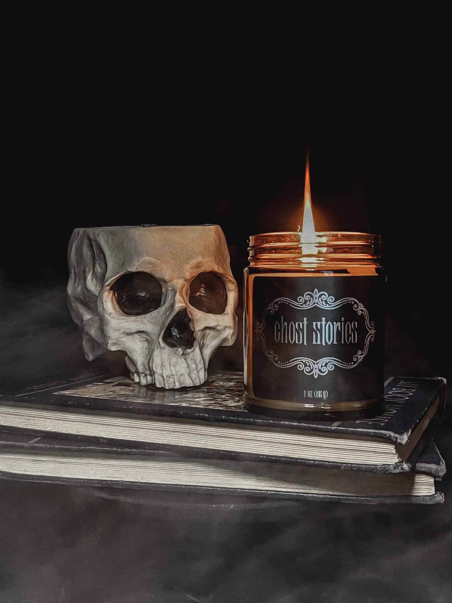 Halloween amber glass jar candle with black label embellished with metallic filigree and the text "Ghost Stories". Candle is lit and sitting on top of two hardback books with black covers. Beside the candle is a creepy grey colored skull with black eye sockets. Top of the skull is open. 