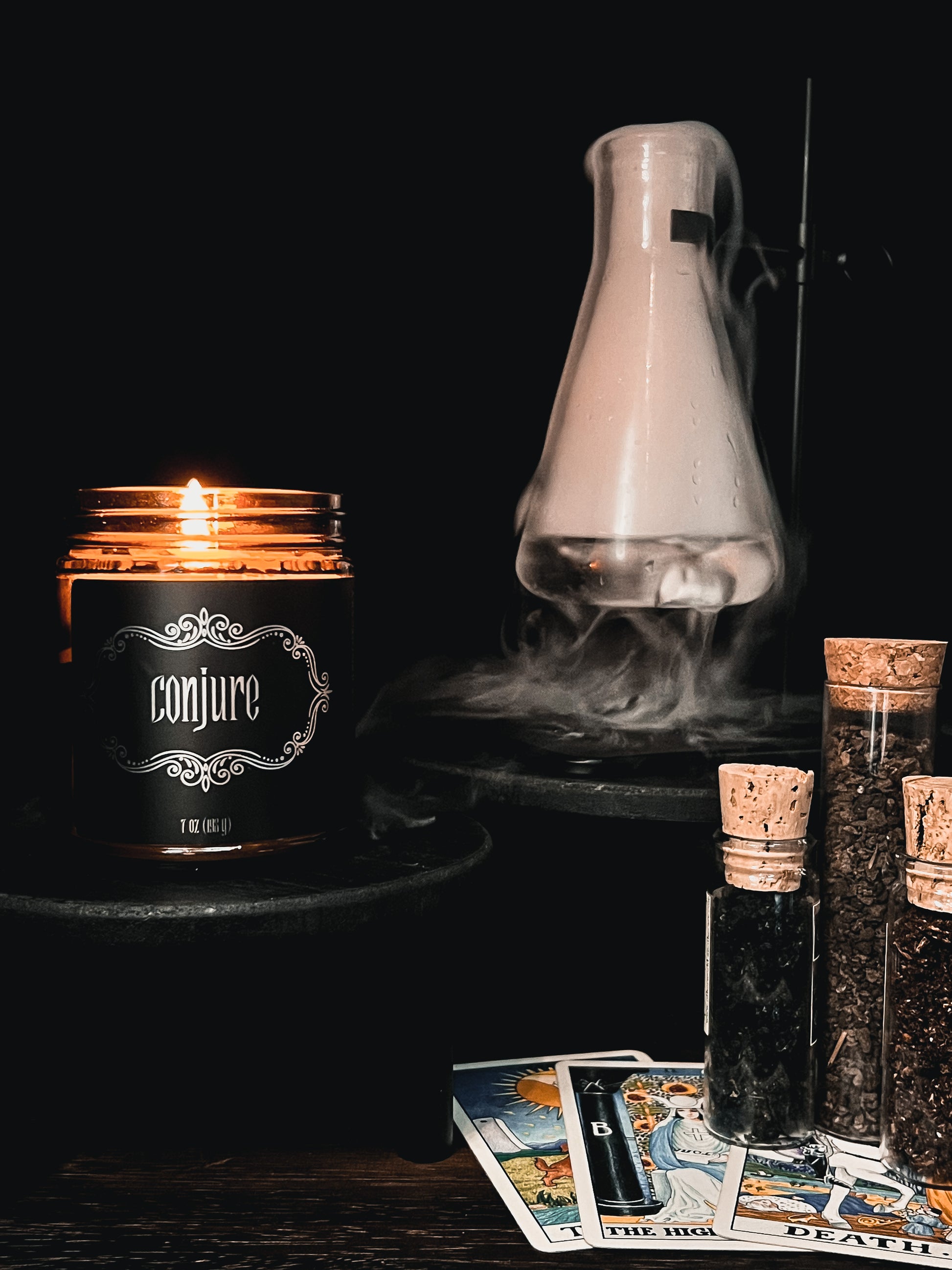 Halloween amber glass jar candle with black label embellished with metallic filigree and the text "Conjure". Surrounded with bubbling beaker and tarot cards showing the Death card.