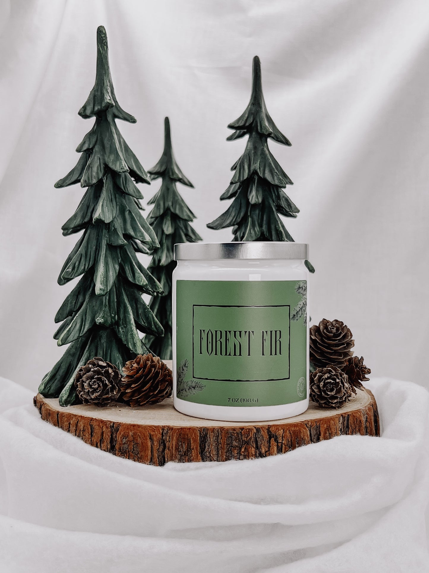 A white jar candle with a silver metal lid and a green metallic matte style label with images of fir tree branches with the text "FOREST FIR" printed on it. The candle sits on top of a wooden disc placed along side it are many small pine cones. Behind the candle are three dark green colored trees.