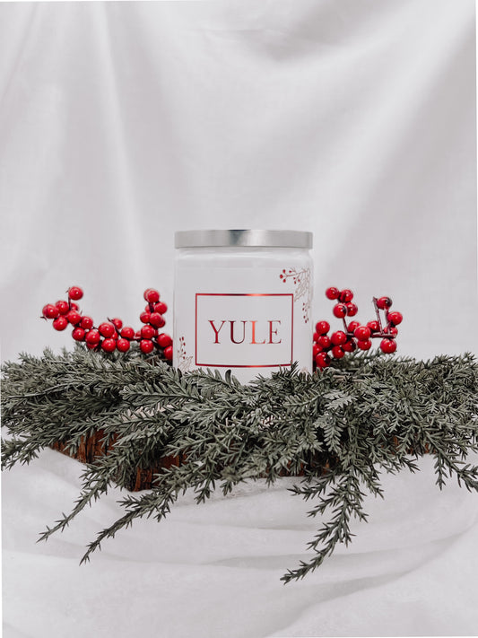 A white jar candle with a silver metal lid and a white metallic label with images of red berries attached to a branch with the text "Yule" printed in red. Candle sits on top of one medium sized tree stump disc surrounded by evergreen branches and red berries. Background is a white back drop.