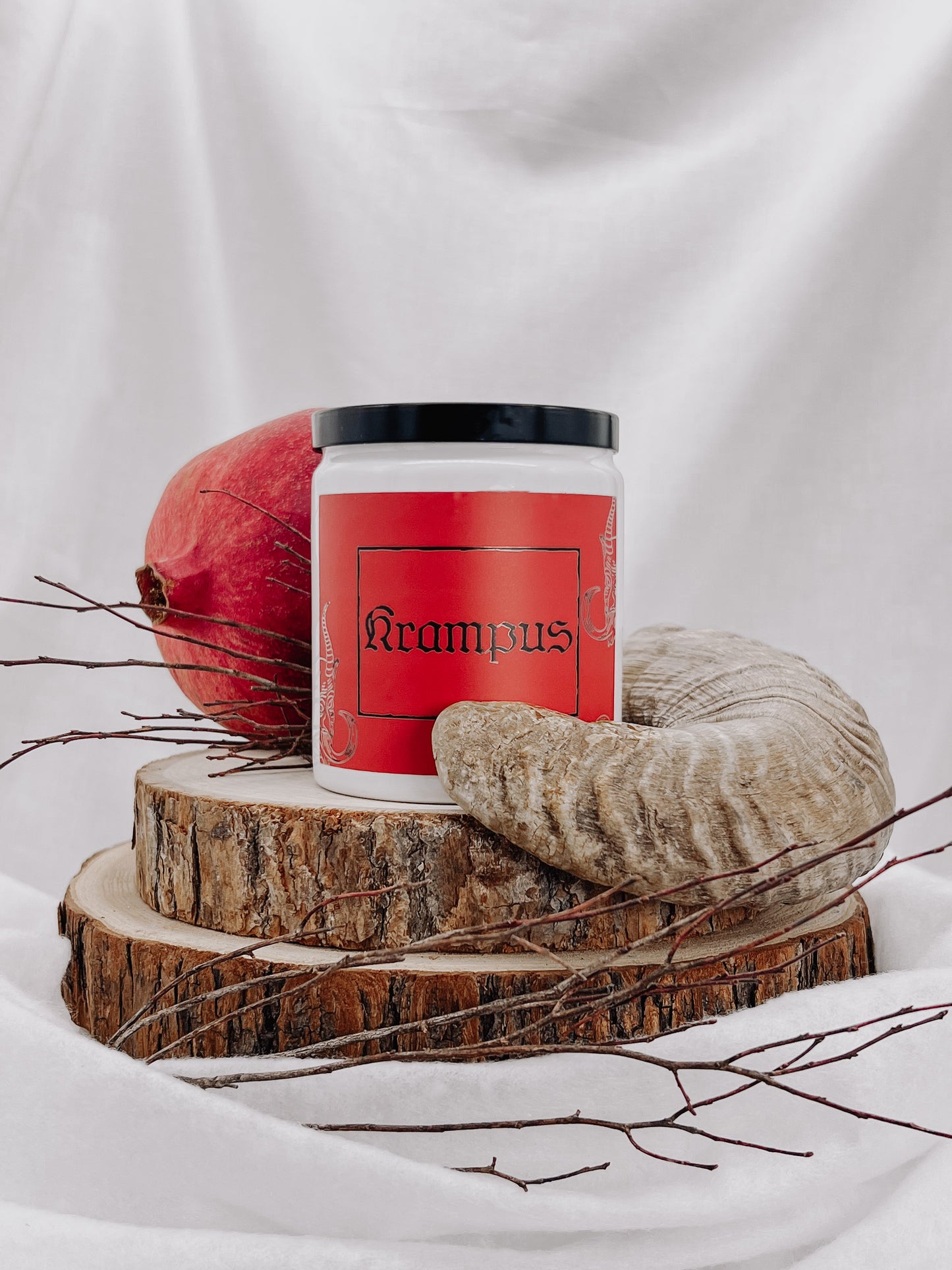 A white jar candle with a black metal lid and a red metallic matte style label with images of krampus's face with the text "Krampus" printed in black. Candle sits on top of a wooden disc placed along side a ram's horn, and a pomegranate, placed on top of two medium sized tree stump discs.