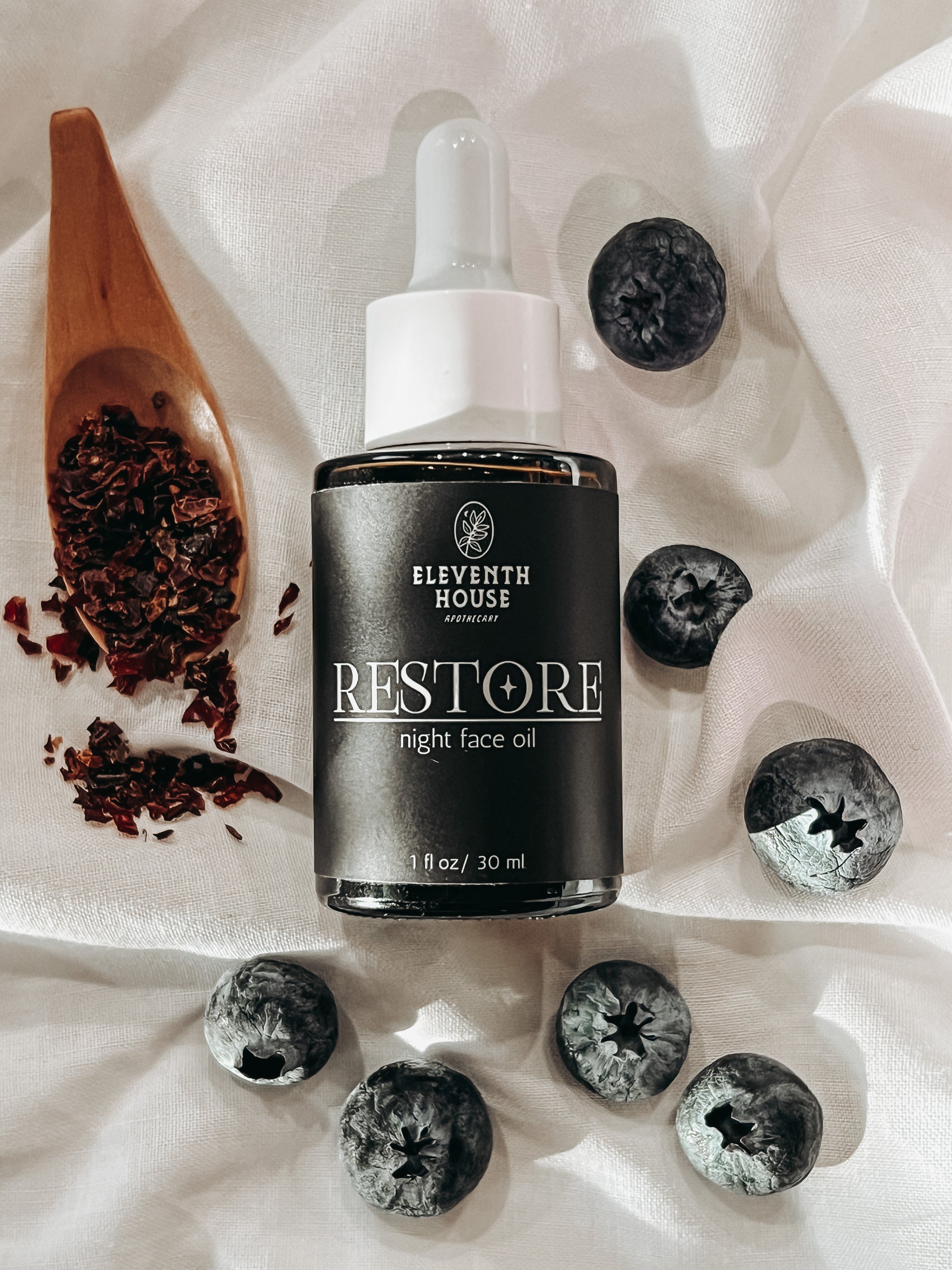 One ounce amber glass dropper bottle with white top and black matte style label with the text "RESTORE night face oil" and Eleventh House Apothecary logo in white lettering. Bottle is laying on it's side on top a white background with fresh blueberries placed to the right and bottom of it. To the left there is a wooden spoon filled with dried rose hips.